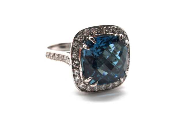 Checkerboard faceted cushion cut London blue topaz claw set above a halo of diamonds on a white gold band